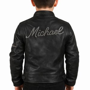 Może przedstawiać: Czarna skórzana kurtka z napisem "Michael" wysadzanym srebrnymi ćwiekami na plecach. Kurtka ma klasyczny styl motocyklowy z zapięciem na zamek błyskawiczny, zatrzaskami i pikowanymi detalami na ramionach. Osoba ma na sobie czarne dżinsy.