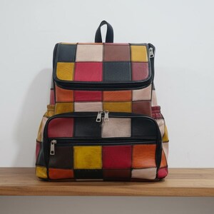 Peut inclure: Sac &agrave; dos patchwork multicolore en cuir avec rabat sup&eacute;rieur et poche avant zipp&eacute;e. Le sac pr&eacute;sente des carr&eacute;s rouges, jaunes, noirs et beiges. Le sac &agrave; dos est pos&eacute; sur une surface en bois.