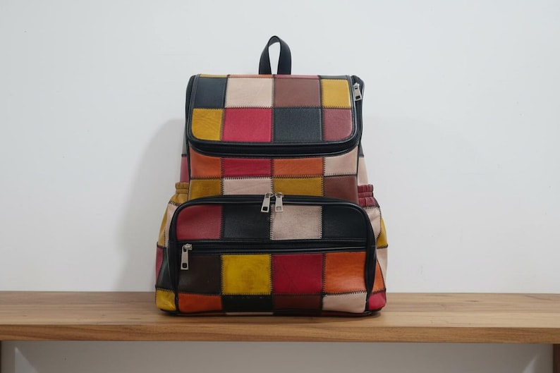 Peut inclure: Sac &agrave; dos patchwork multicolore en cuir avec rabat sup&eacute;rieur et poche avant zipp&eacute;e. Le sac pr&eacute;sente des carr&eacute;s rouges, jaunes, noirs et beiges. Le sac &agrave; dos est pos&eacute; sur une surface en bois.