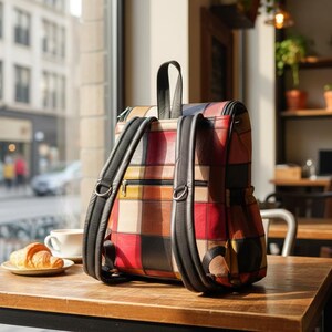 Peut inclure: Un sac &agrave; dos patchwork multicolore avec des bretelles et une poign&eacute;e noires. Le sac est pos&eacute; sur une table en bois, avec une tasse de caf&eacute; et un croissant. Le design du sac comprend des carr&eacute;s rouges, bruns et beiges.