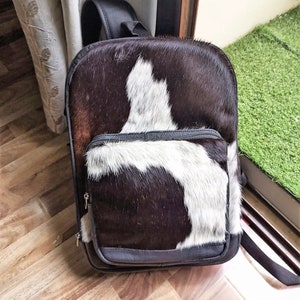 Puede incluir: Una mochila de piel de vaca negra y blanca con un bolsillo con cremallera en la parte delantera. La mochila tiene un ribete de cuero marrón y correas ajustables.