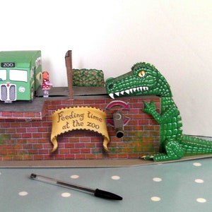 Könnte beinhalten: Eine skurrile Papierszene eines Zoos mit einem grünen Bus, einer Backsteinmauer mit dem Schild "Feeding time at the zoo" und einem großen grünen Alligator. Ein Stift liegt im Vordergrund.