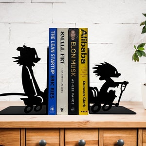 May include: Black metal bookends shaped like cartoon characters riding scooters. The bookends are holding up a stack of books with titles like "Alibaba", "Elon Musk", and "The Lean Startup".