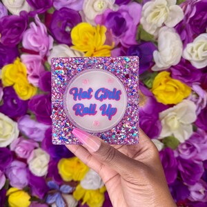 Puede incluir: Cenicero cuadrado con purpurina y el texto "Hot Girls Roll Up" en rosa y azul. El cenicero se sostiene frente a un fondo de flores moradas, blancas y amarillas.