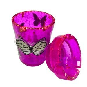 May include: A pink and gold glitter ashtray with a butterfly design. The ashtray is made of resin and has a removable lid.
