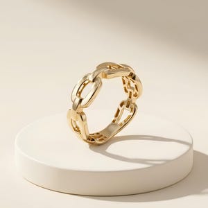 May include: A polished gold ring featuring a continuous chain link design. The band is composed of interconnected oval-shaped metal links with a smooth, reflective surface, creating a uniform and repeating pattern around the entire circumference.