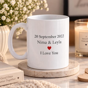 May include: A white ceramic mug featuring the black printed text 20 September 2022, Nima & Leyla, a small red heart icon, and the text I Love You. The mug has a smooth finish and a curved handle, presented in a clean, minimalist style.