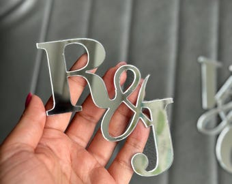 May include: A set of mirrored acrylic letters spelling out "R & J". The letters are held in a hand, with a blurred background. The letters are reflective and appear to be silver.
