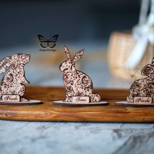 May include: Three wooden Easter bunny figurines with detailed floral patterns, each with a nameplate. The bunnies are on a wooden tray, with a basket in the background. The names on the nameplates are Noah, Jane, and Nicole.