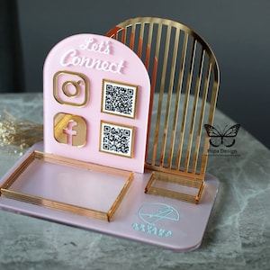 May include: Pink acrylic desk organiser with gold accents. It features a 'Let's Connect' sign with Instagram and Facebook icons, two QR codes, and a small tray. The organiser has a gold arch design and a butterfly logo with the text 'Pupa Design'.
