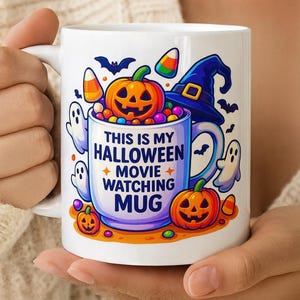 Op de afbeelding: Witte keramische Halloween-mok met een handvat en een kleurrijk ontwerp. De mok heeft een heksenhoed, pompoenen, geesten, snoepmaïs en de tekst "THIS IS MY HALLOWEEN MOVIE WATCHING MUG". De mok wordt door een persoon vastgehouden.