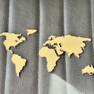 May include: Gold-coloured world map cutouts on a grey, textured surface. The continents are clearly defined, including North America, South America, Europe, Africa, Asia, and Australia. These cutouts are likely decorative items.