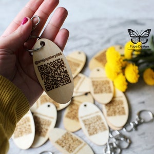 Può includere: Portachiavi in legno con un codice QR inciso a laser e il testo "@artejegellery888". I portachiavi sono di forma ovale e hanno un anello di metallo argentato. I portachiavi sono esposti su una superficie bianca con fiori gialli.