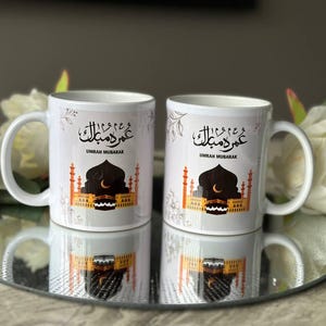 Könnte beinhalten: Zwei weiße Keramikbecher mit schwarzer arabischer Kalligraphie, die "Umrah Mubarak" lautet, und einer Darstellung der Kaaba. Die Becher haben ein florales Design und stehen auf einem weißen Hintergrund.