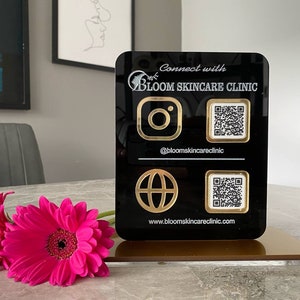 May include: Black sign with gold lettering that says "Connect with Bloom Skincare Clinic". The sign has a gold Instagram icon, a QR code, a gold globe icon, and another QR code. The sign is on a gold stand.