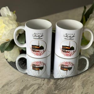May include: Two white ceramic mugs featuring black Arabic calligraphy that reads "Eid Mubarak" and a graphic of the Kaaba in Mecca. The mugs have pink floral accents and are sitting on a white and silver coaster.
