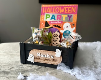 Puede incluir: Una caja de madera negra llena de golosinas de Halloween y un libro de actividades. La caja presenta un letrero de madera que dice "Noah's Halloween Sweets & Treats" con recortes de fantasmas. El libro de actividades se titula "Halloween Party".