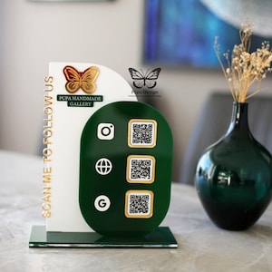 May include: A white and green acrylic display stand with a gold butterfly and the text "Pupa Handmade Gallery" and "Scan Me To Follow Us". The stand has three QR codes and icons for Instagram, Google, and a globe.