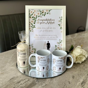 Puede incluir: Un juego de regalo de bodas con dos tazas blancas, una impresión enmarcada y una vela. Las tazas y la vela tienen un diseño floral y el texto "Congratulations on your Nikkah". La impresión enmarcada tiene el mismo texto y una ilustración de pareja de bodas.