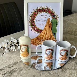 May include: An engagement gift set comprising a framed illustration, two white mugs, and a candle. The illustration depicts a couple with the text "Congrats on your engagement". The mugs and candle share a matching design.