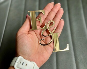 May include: Gold-colored metal letters "F & L" are held in a hand. The letters are in a script font and have a reflective surface. The hand is wearing a white watch with a white band.