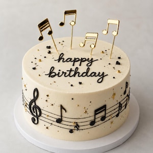 Puede incluir: Un pastel redondo con glaseado suave de color blanco roto, con el texto happy birthday en cursiva negra. Los lados están decorados con notas musicales y pentagramas negros. Adornos de notas musicales doradas y pequeñas chispas doradas decoran la parte superior.