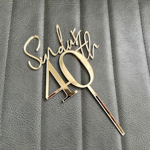 May include: A gold cake topper with the name "Synduth" and the number "40". The topper is made of a reflective material and is designed to be inserted into a cake. The background is a textured grey surface.