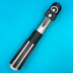 Op de afbeelding: Een zilveren en zwart lichtzwaard rekwisiet met een zwarte handgreep en een zwarte bovenkant met een wit halve cirkel ontwerp. Het lichtzwaard heeft een cilindrisch lichaam met een zwarte grip, tegen een helderblauwe achtergrond.