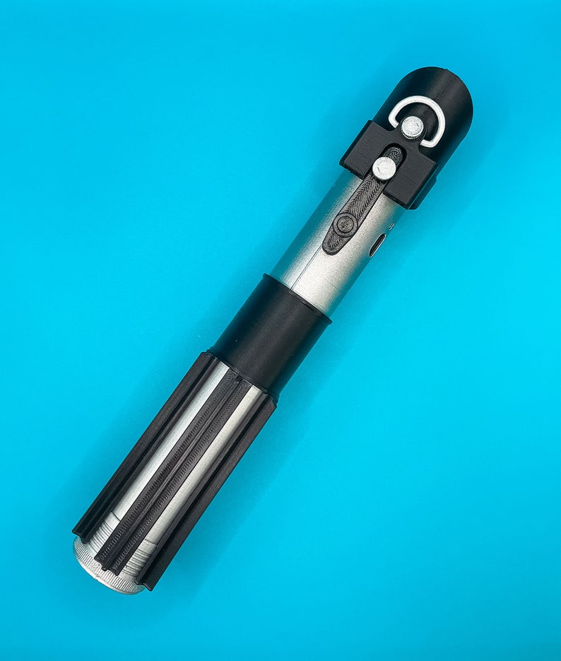 Op de afbeelding: Een zilveren en zwart lichtzwaard rekwisiet met een zwarte handgreep en een zwarte bovenkant met een wit halve cirkel ontwerp. Het lichtzwaard heeft een cilindrisch lichaam met een zwarte grip, tegen een helderblauwe achtergrond.