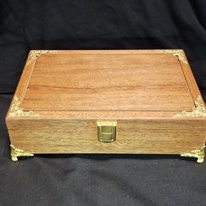 May include: A rectangular wooden box with a light brown finish. The box has gold-coloured corner accents, a clasp, and decorative feet. The box is closed and sits on a black surface.