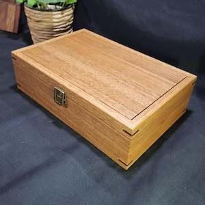May include: A rectangular wooden box with a natural light brown grain, featuring a hinged lid and a small brass-coloured metal latch on the front. The corners of the box are constructed with visible dark wood splines for reinforcement and detail.