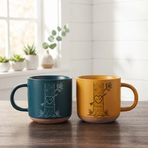 May include: Two ceramic mugs, one dark teal and one mustard yellow, each featuring a black line art illustration of a tree trunk with a heart carved into the bark. The heart contains the letters L+A and an arrow, with the date 17.05.26 below.