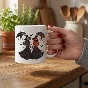 Puede incluir: Una taza de cerámica blanca con una ilustración gráfica de dos figuras con vestidos largos negros y sombreros puntiagudos, cada una sosteniendo un paraguas negro. Las figuras tienen el pelo largo y están rodeadas de pequeños murciélagos negros.