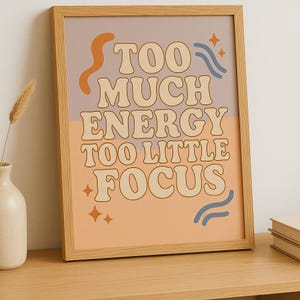 May include: A framed print with the text "TOO MUCH ENERGY TOO LITTLE FOCUS" in a retro font. The print has a color scheme of peach, lavender, and beige, with orange and blue decorative elements. The frame is made of light-colored wood.