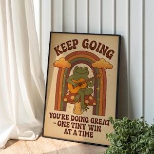 Puede incluir: Un póster enmarcado con un diseño retro. Presenta una rana tocando la guitarra sobre una seta, con un arcoíris y las palabras "KEEP GOING" y "YOU'RE DOING GREAT - ONE TINY WIN AT A TIME". La paleta de colores incluye marrones, naranjas y verdes.