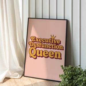 Könnte beinhalten: Ein gerahmtes rosa Poster mit den Worten "Executive Dysfunction Queen" in einer Retro-Schriftart. Der Text ist in Gelb- und Marontönen gehalten, mit einer kleinen Krone über dem Wort "Queen". Das Poster wird vor einer weißen Wand ausgestellt.