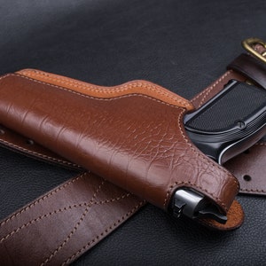 May include: A brown leather holster with a black handgun inside. The holster is attached to a brown leather belt with a metal buckle.