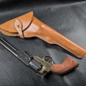Könnte beinhalten: Ein schwarzer Metallrevolver mit Messingrahmen und Holzgriff, der neben einem beigefarbenen Lederholster liegt. Das Holster verfügt über einen Klappverschluss, der mit einem Messingknopf gesichert ist. Der Revolver hat einen langen Lauf.