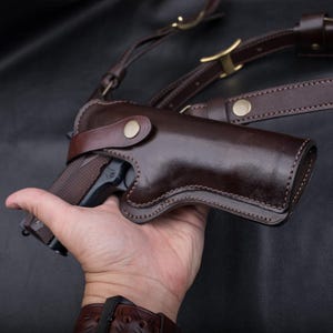 Può includere: Una fondina da spalla in pelle marrone scuro con finitura lucida, chiusura con bottone a pressione e cinghie in pelle regolabili con fibbie in metallo color ottone, progettata per sostenere una pistola in posizione orizzontale.