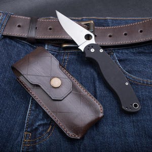 Può includere: Un fodero per coltello in pelle marrone con un motivo a rombi cucito e un coltellino tascabile nero con una lama argentata. Il coltello e il fodero sono su un paio di jeans blu con una cintura in pelle marrone.