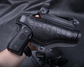 May include: A black leather holster with a crocodile-embossed design. The holster is designed to hold a handgun and has a strap for carrying.
