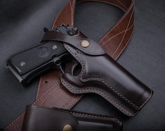 May include: A brown leather holster with a black handgun inside. The holster is attached to a brown leather belt.