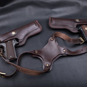 Könnte beinhalten: Ein doppeltes Schulterholster-System aus dunkelbraunem Leder mit zwei Holstern für halbautomatische Pistolen. Das System umfasst verstellbare Riemen mit messingfarbenen Metallschnallen und Nieten sowie ein zentrales Rückenteil mit Prägung.