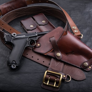 May include: A black semi-automatic pistol with a textured grip rests on a brown leather holster and a matching leather belt. The holster features a flap with a brass snap closure, and the belt includes a large brass buckle and metal hardware.