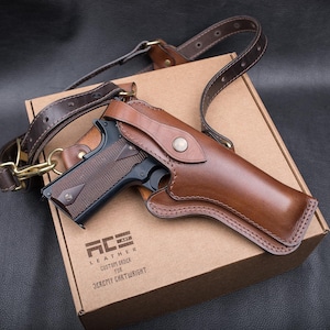 Könnte beinhalten: Ein braunes Schulterholster aus Leder mit einer schwarzen halbautomatischen Pistole und einem passenden Lederriemen mit Messingbeschlägen. Das Holster liegt auf einem Karton mit dem Aufdruck ACE LEATHER CUSTOM STOCK FOR JEREMY CARTWRIGHT.