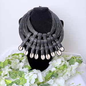 Op de afbeelding: Een zwarte statement ketting op een zwarte mannequin. De ketting heeft meerdere strengen zwart koord, geweven secties en decoratieve kaurischelpen. Witte bloemen en groene bladeren omringen de ketting.
