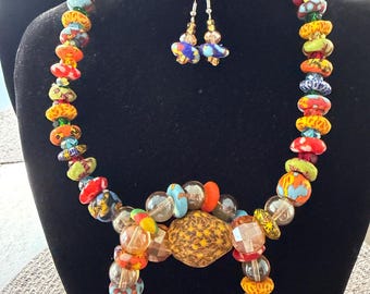 May include: A vibrant necklace and earring set with multi-coloured glass beads. The necklace includes red, orange, yellow, blue, and clear beads. The earrings mirror the necklace's design. The jewellery is presented on a black mannequin.