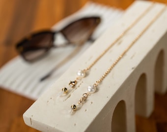 May include: Two gold eyeglass chains with pearl accents. The chains are displayed on a white surface.