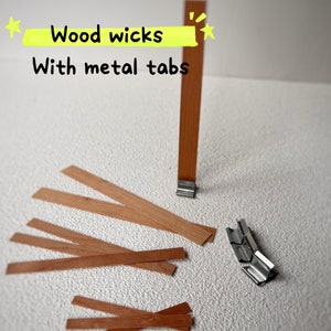 May include: A close-up of wooden wicks with metal tabs. The wicks are brown and the tabs are silver. The text "Wood wicks with metal tabs" is written in yellow on a white background.