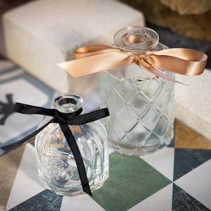 May include: Two clear glass bottles with decorative designs, tied with ribbons. The larger bottle has a gold ribbon, and the smaller bottle has a black ribbon.